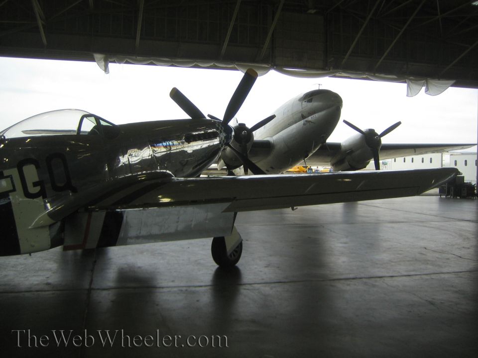 Polished silver fighter aircraft marked GQ parked beneath wing of larger plane in hangar