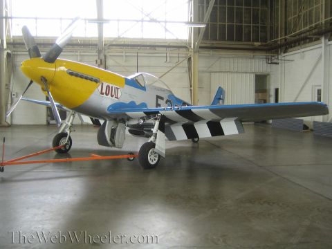 1990 Ford Bronco - silver fighter aircraft with a yellow nose, blue trim and black-checkered tail, marked 'LOU IV', sits inside a hangar.