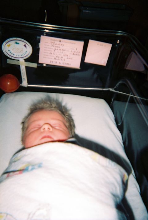 2002 Oldsmobile Aurora - A newborn baby rests peacefully in a hospital bassinet, adorned with a safety sticker and a card detailing birth information.