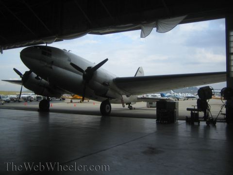 1990 Ford Bronco - silver twin-engine propeller transport aircraft sits in an open hangar with film lighting equipment nearby and planes on the ramp beyond.