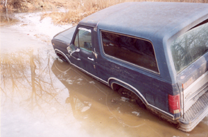 1982 Ford Bronco - A dark blue SUV is stuck in muddy water, with its tires submerged.