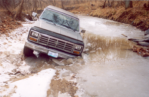 1982 Ford Bronco - A Ford Bronco is stuck in a muddy, watery ditch, with its front tires deep in the muck.