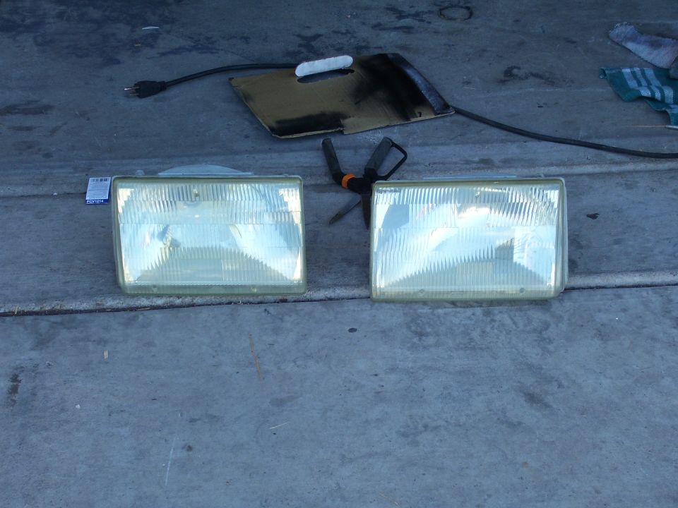 Two rectangular headlights with clear lenses and ribbed interiors sit on a concrete surface next to tools