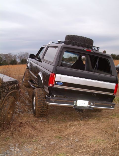 1995 Ford Bronco - Rear three-quarter view of a lifted black Ford Bronco tipped over a rocky rut in a dry grass field, tailgate glass rolled down to show the…
