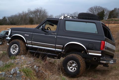 1995 Ford Bronco - Black lifted Ford Bronco parked at an angle on a rocky, grassy embankment, seen from the driver's side in three-quarter rear view with the…