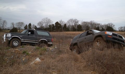 1995 Ford Bronco - Two SUVs sitting nose-down on a brushy, rocky field on an overcast winter day: a black lifted Ford Bronco with chrome trim and a roof rack…