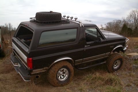 1995 Ford Bronco - Elevated three-quarter rear view of a black lifted Ford Bronco parked on rough grass and gravel, showing the removable hardtop, roof rack w…