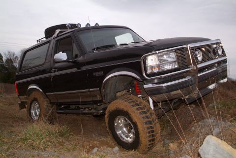1995 Ford Bronco - Close low front three-quarter view of a lifted black Ford Bronco flexed over a rocky ledge in dry grass, front tire caked in mud and a red…
