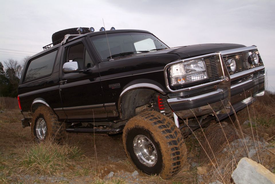 Lifted black Ford Bronco flexed over rocks, close front 3/4 view with muddy tire