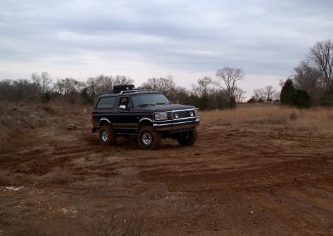 1995 Ford Bronco - Lifted black Ford Bronco sitting in a churned-up dirt field with mud packed on its tires and rocker panels, seen from a distant front three…