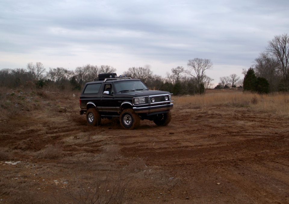 Muddy lifted black Ford Bronco in a torn-up dirt field under overcast sky