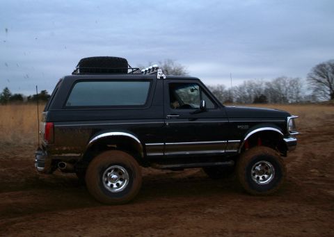 1995 Ford Bronco - Black lifted Ford Bronco being driven across a muddy dirt field, shot from the passenger side in profile with a driver visible through the…