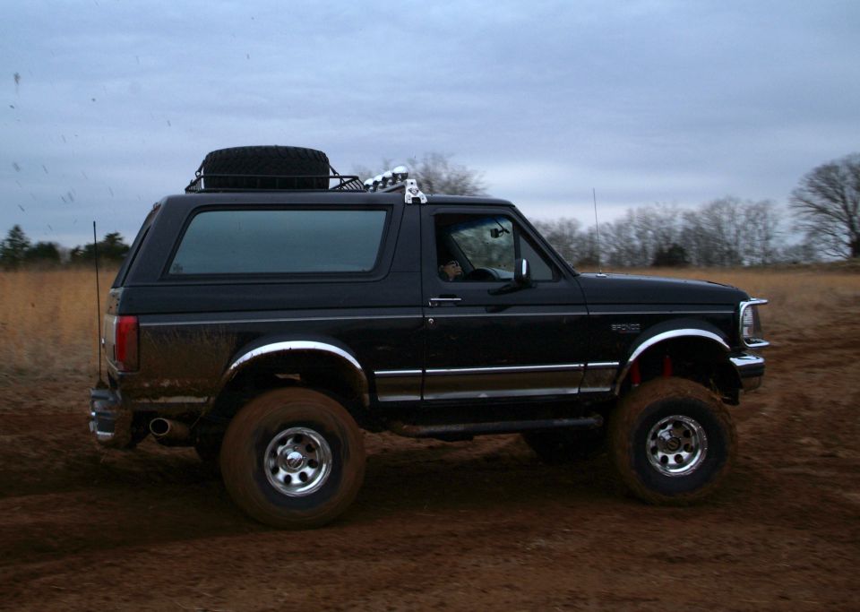 Black lifted Ford Bronco driving through muddy field, side profile
