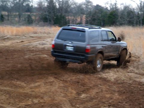 1995 Ford Bronco - SUV driving through a muddy field with trees in the background.