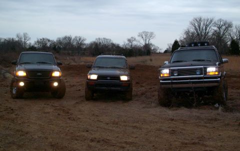 1995 Ford Bronco - Three SUVs, including a 1995 Ford Bronco, are parked on a dirt lot under an overcast sky.