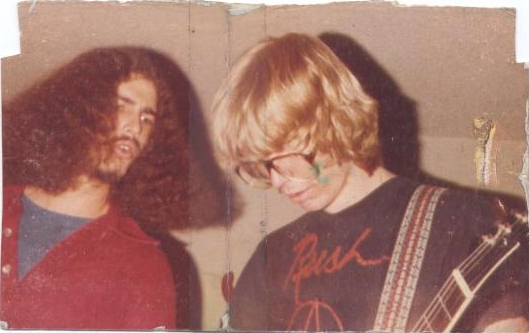 Two men posing for a photo, one with long dark hair and a red jacket, the other with blond hair and glasses holding a guitar