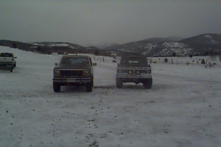 Two trucks facing forward parked on snowy ground with hills behind under gray sky