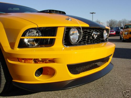 Yellow Ford Mustang front end with black stripes and hood scoop. Another yellow car is visible in the background