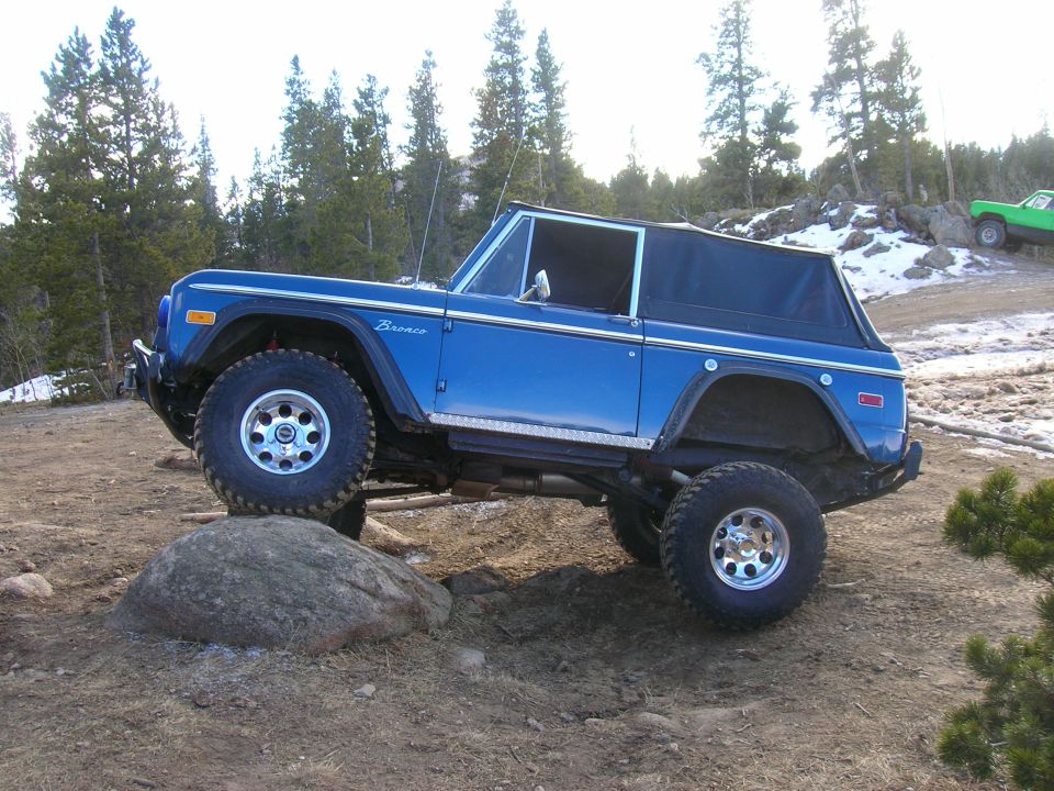 Blue Ford Bronco with lifted suspension driving over a rock on a dirt trail. Another green vehicle is visible on a hill in the background