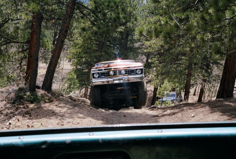 Brown off-road vehicle with lifted suspension and large tires, front view, driving on a dirt path between trees. Another vehicle is visible in the background