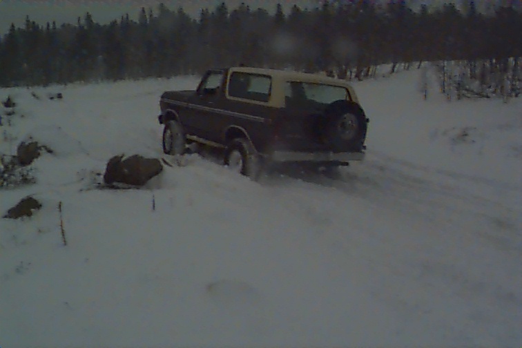 Dark brown and tan Ford Bronco driving in snow, spare tire on rear