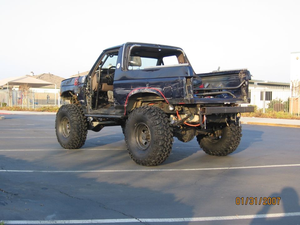 Dark blue lifted Ford Bronco, stripped body, big mud tires, rear 3/4 view in parking lot