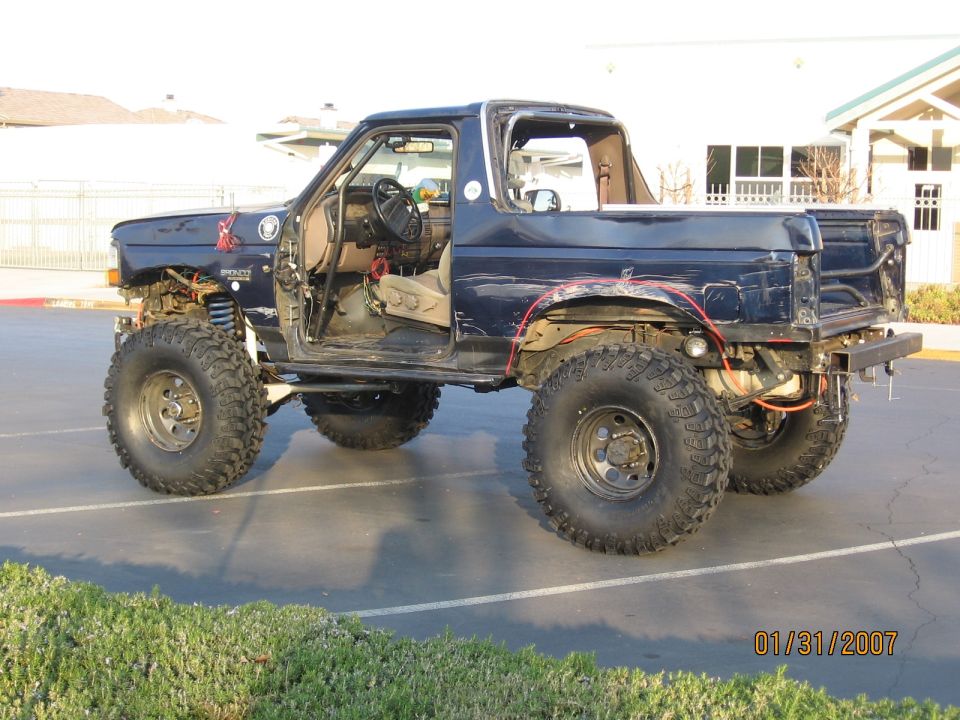 Lifted dark blue Ford Bronco on big tires, doors removed, side/rear view in parking lot
