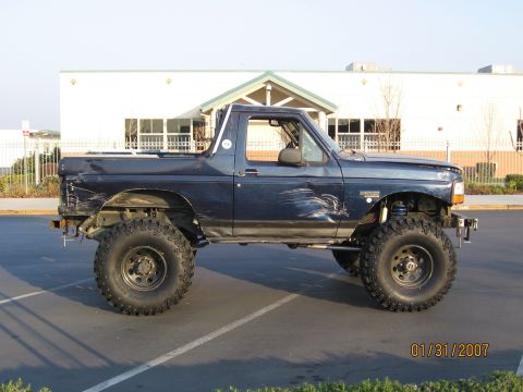 1994 Ford Bronco - Modified Ford Bronco with off-road tires and visible body damage sits parked on asphalt.