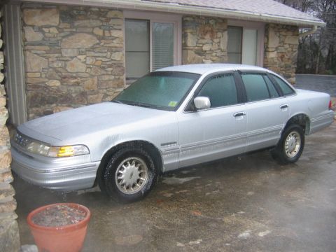 1985 Ford Crown Victoria LTD - Silver sedan parked outside a stone house on a wet day.