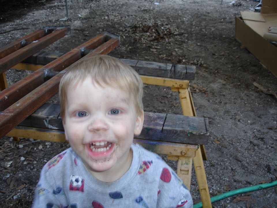 Child with dirty face and a smile, wearing a grey shirt with soccer ball graphics. Wooden structures and dirt ground in background