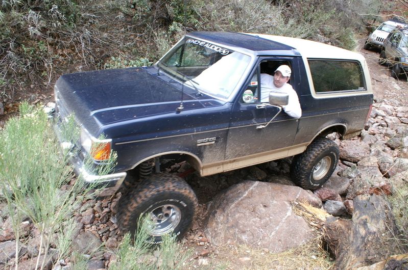 Dark blue Ford Bronco with white top, driver in white hat, navigating rocky terrain. Another SUV is visible behind it