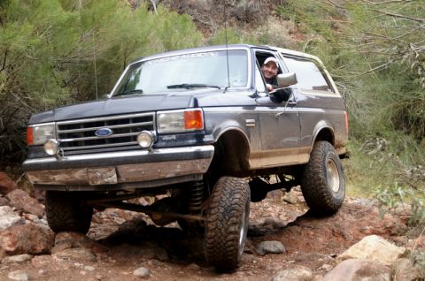 1991 Ford Bronco - A man smiles from the driver's seat of a dark blue Ford Bronco navigating a rough, off-road path.