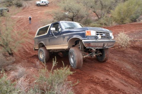 1991 Ford Bronco - A dark blue 1991 Ford Bronco with a white top navigates a steep, dusty dirt trail, with another vehicle visible further up the incline.