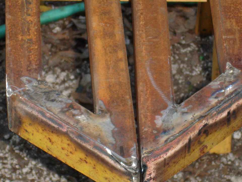 Close-up of rusty metal bars welded together, with a green hose and gravel in the background. Supermotors.net logo visible