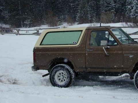 1980 Ford Bronco - A brown 1980 Ford Bronco is seen from the side, driving through a snowy landscape with trees in the background.