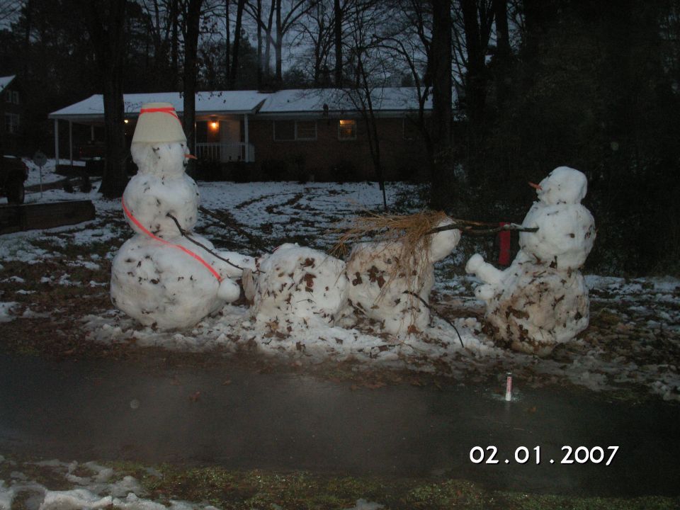 Three snowmen in a yard with a house in the background; one snowman has a bucket on its head