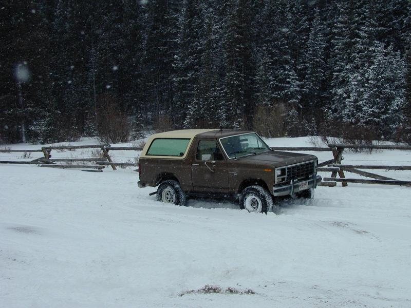 Brown Ford Bronco parked in deep snow with trees and a wooden fence in the background