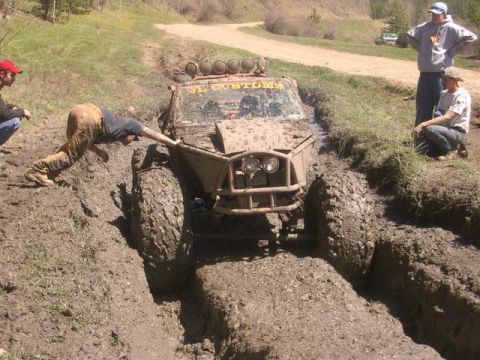1995 Ford Bronco - An off-road vehicle is deeply stuck in a muddy rut, with several people attempting to free it.