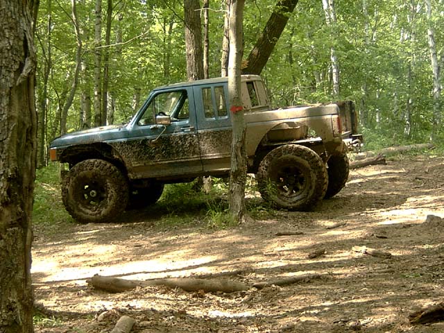 Muddy dark green lifted Ford pickup parked on a forest dirt trail, side view.