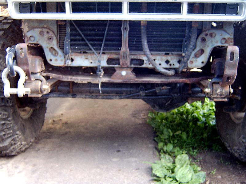 Underside front view of truck showing rusty crossmember, radiator, frame horns, white shackle, and off-road tires.