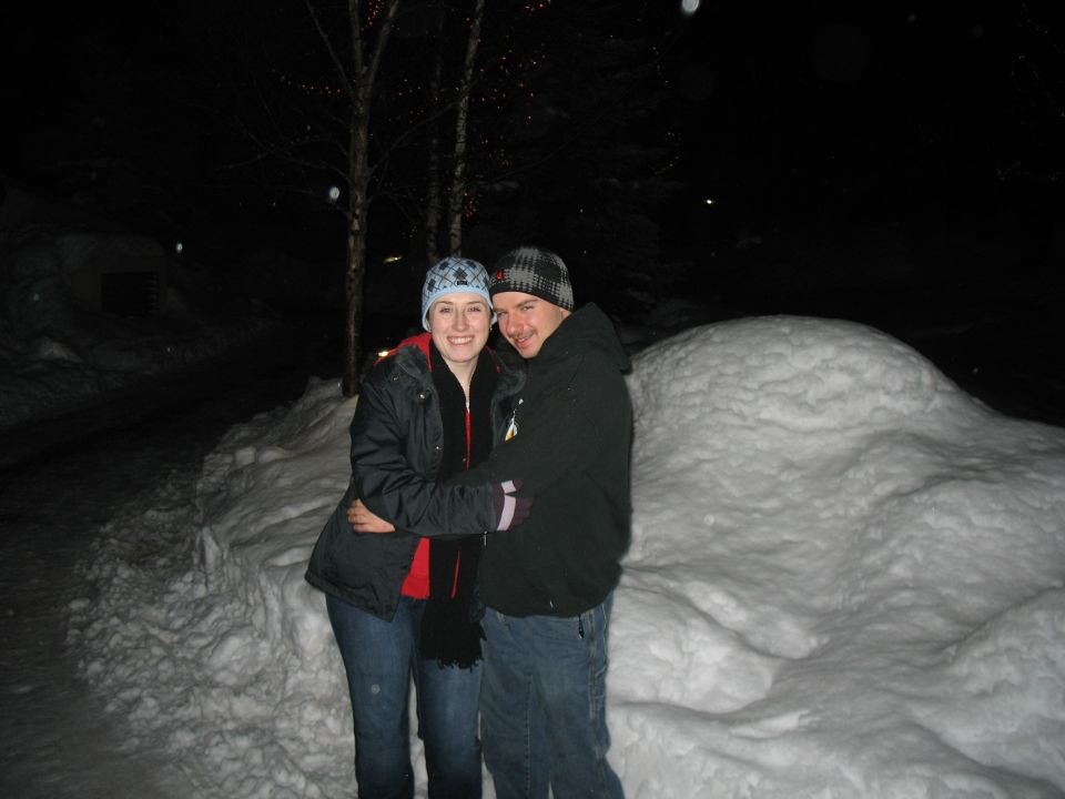 Couple hugging in the snow at night, with trees and lights in the background