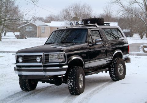 1995 Ford Bronco - Front three-quarter view of a lifted black Ford Bronco on a snow-dusted driveway, wearing a chrome brush guard and bumper, chrome fender fl…
