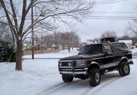 1995 Ford Bronco - Wide front three-quarter shot of a lifted black Ford Bronco parked on a snow-covered residential street, tire tracks cut in the snow behind…
