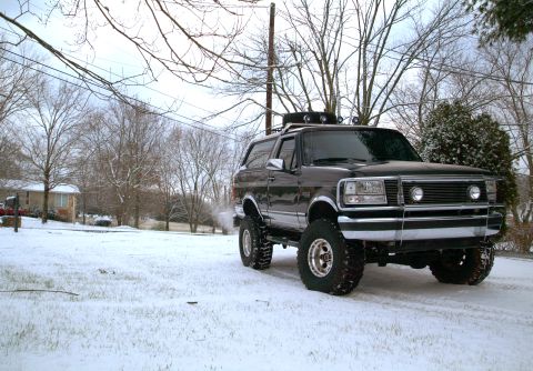 1995 Ford Bronco - Black lifted Ford Bronco parked in a snowy residential yard, shot from the driver's side in three-quarter front view with exhaust vapor dri…