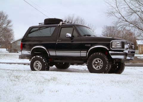 1995 Ford Bronco - Black lifted Ford Bronco full-size SUV parked in a snow-covered yard, shot from the passenger side in three-quarter front view. It wears ch…
