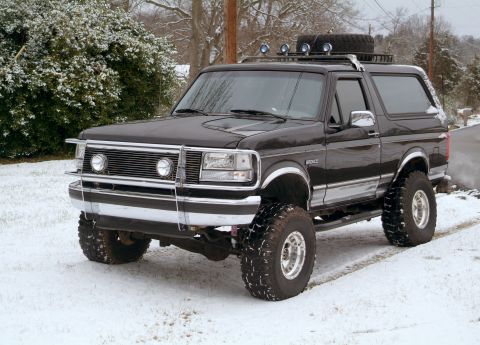 1995 Ford Bronco - Three-quarter front view of a lifted black Ford Bronco parked in fresh snow, wearing a chrome brush guard, chrome bumper, mud-terrain tires…