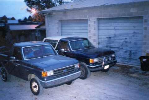 1989 Ford Bronco - Two Ford vehicles, a pickup and a Bronco, are parked outside a garage. The pickup appears to be from the late 1980s.