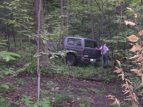 1989 Ford Bronco - A 1989 Ford Bronco is seen partially off-road in a wooded area with a person standing nearby.