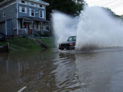 1989 Ford Bronco - A dark SUV navigates through flooded street water, sending up massive sprays on either side.