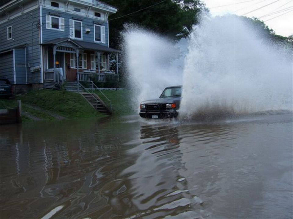 Dark SUV driving through floodwater, creating large splashes. A blue house is visible on the left